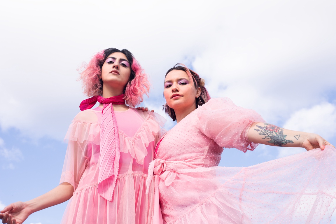 Namra Beatrix Saleem og Sarah Christin Calvert er ikledd rosa, luftige kjoler og ser ned mot fotografen. I bakgrunnen er det blå himmel og store, fluffy skyer.