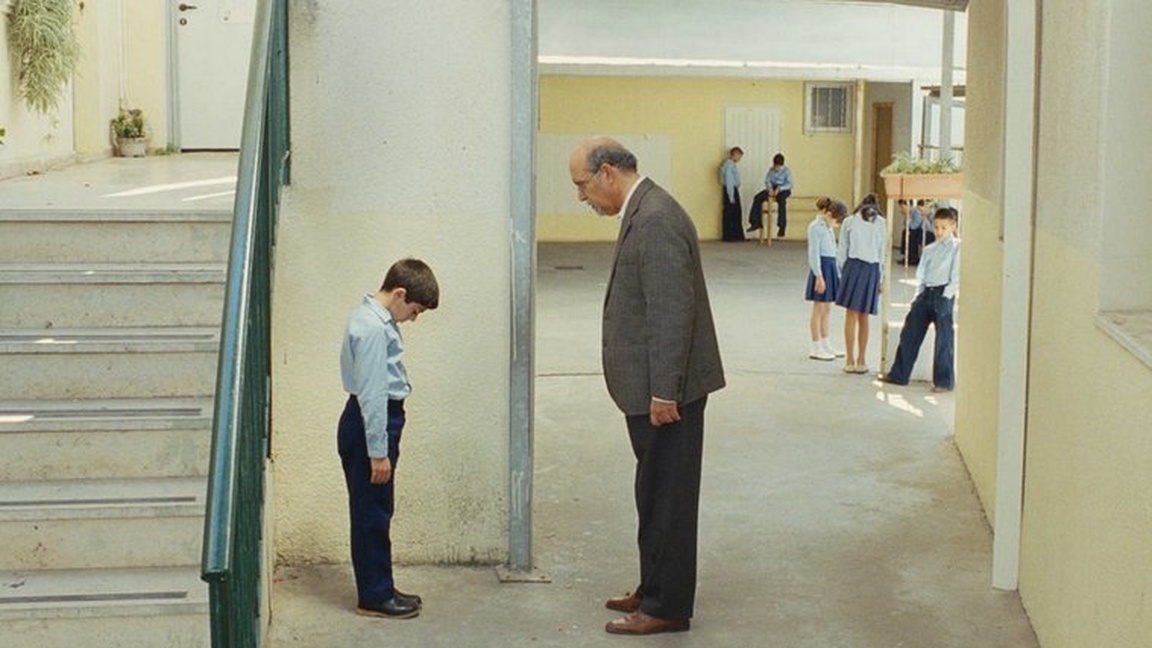 En gutt står med skoleuniform og ser ned i gulvet. Foran han står en voksen mann i dress og ser på ham. Rundt dem ser man en skole, med klasserom og andre barn.