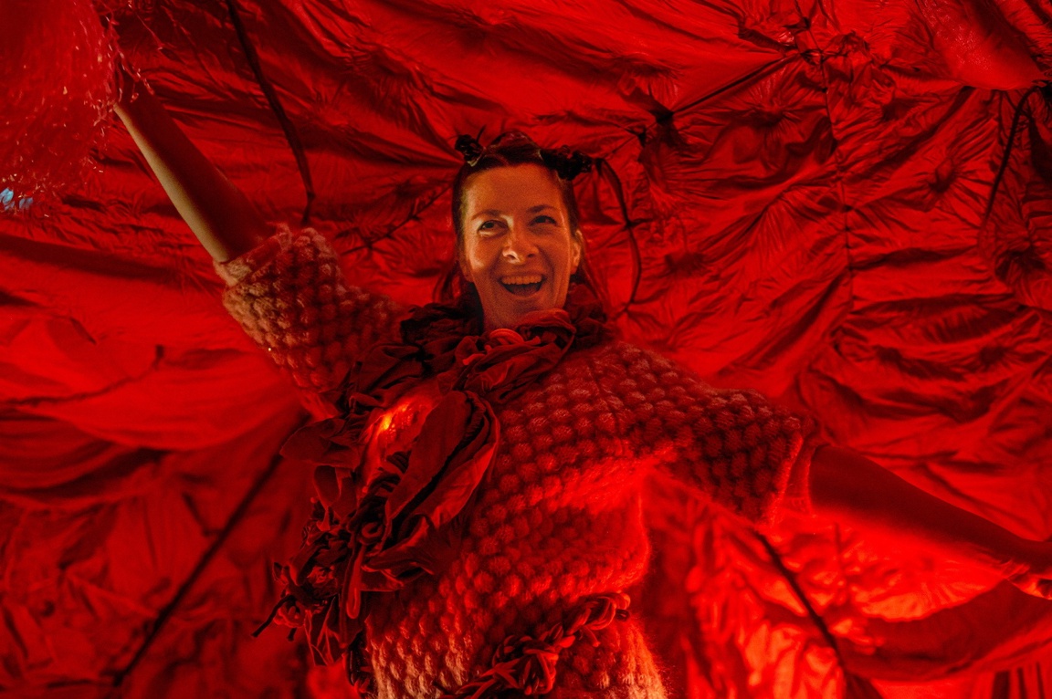 En kvinne kledd i rødt står med strake armer. mot en rød tekstilbakgrunn.