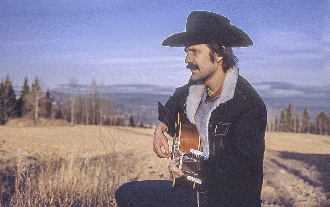 En mann med Cowboyhatt spiller på en gitar. Kulturlandskap i bakgrunnen.